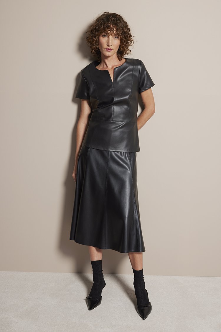 Full-length studio shot of a woman in a black faux-leather top and skirt with matching sheer socks and black pointed-toe heels.