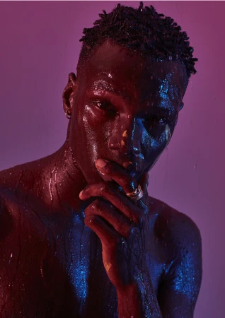 Close-up portrait of a black man with wet skin, illuminated by magenta and blue light, holding hand to face