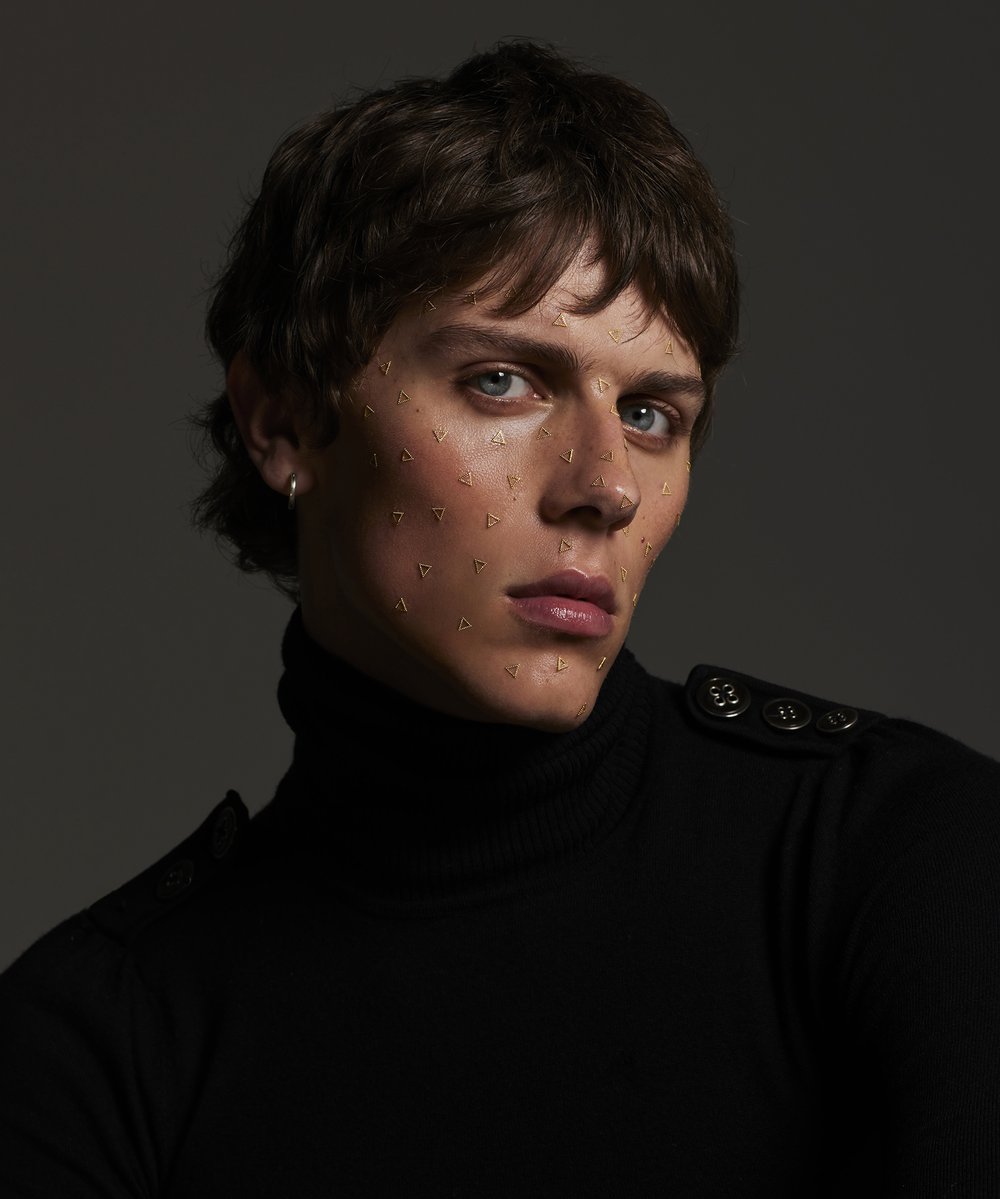 Portrait of a young adult with dark hair wearing a black turtleneck and triangle face gems, posing in front of a grey background.