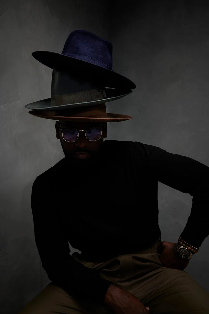 Man wearing glasses and a black turtleneck balancing a stack of three fedoras on his head