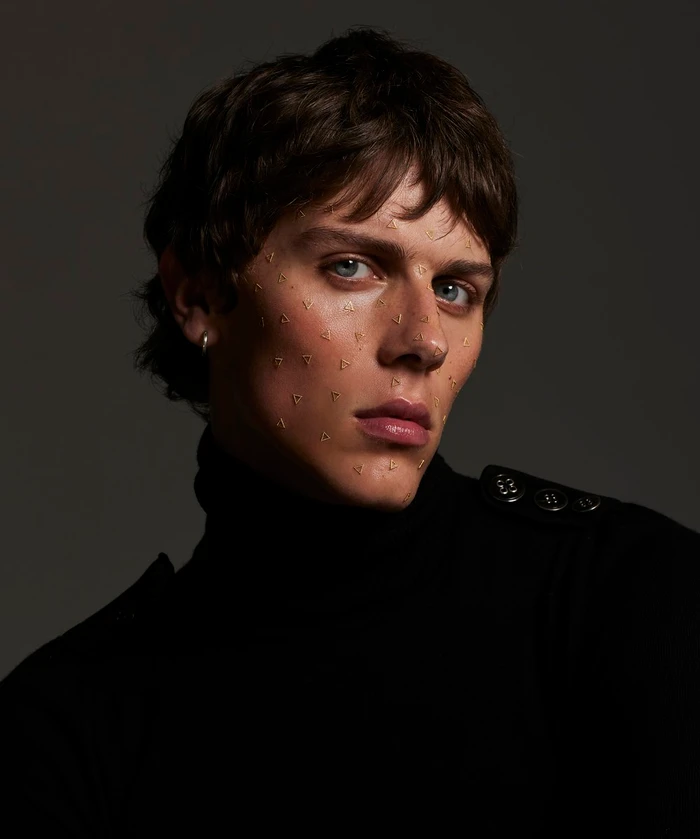 Close-up portrait of a young man wearing a black turtleneck with gold triangle face adornments by Marija Ivkovic