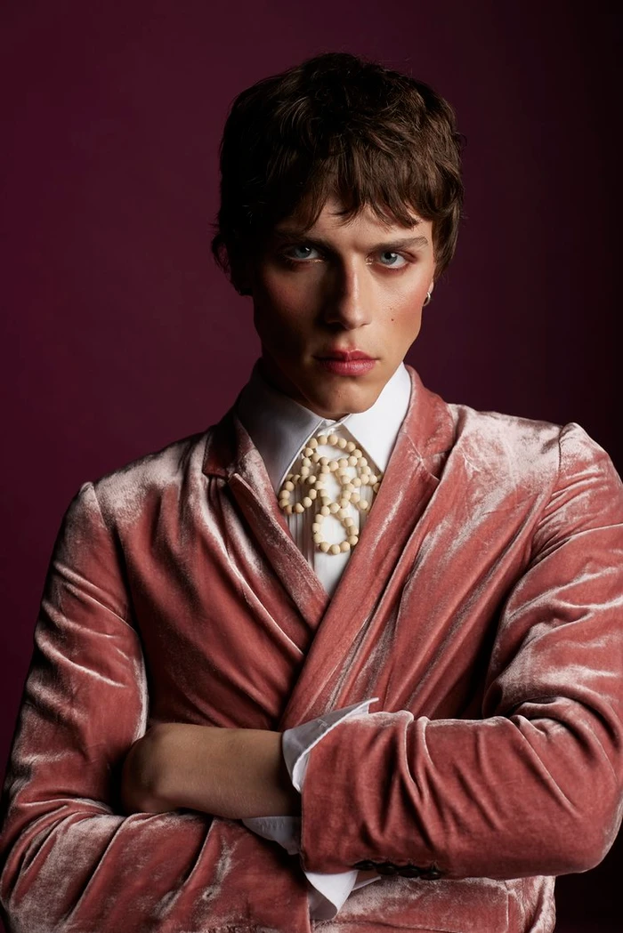 Close-up portrait of a man wearing a dusty pink velvet jacket over a white collared shirt with a beaded neck accessory.