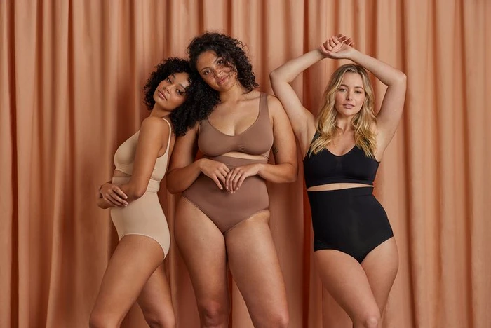 Three female models posing in matching shapewear underwear against a draped peach curtain background.