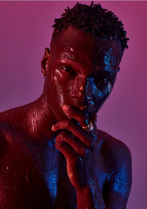 Close-up portrait of a black man with wet skin, illuminated by magenta and blue light, holding hand to face