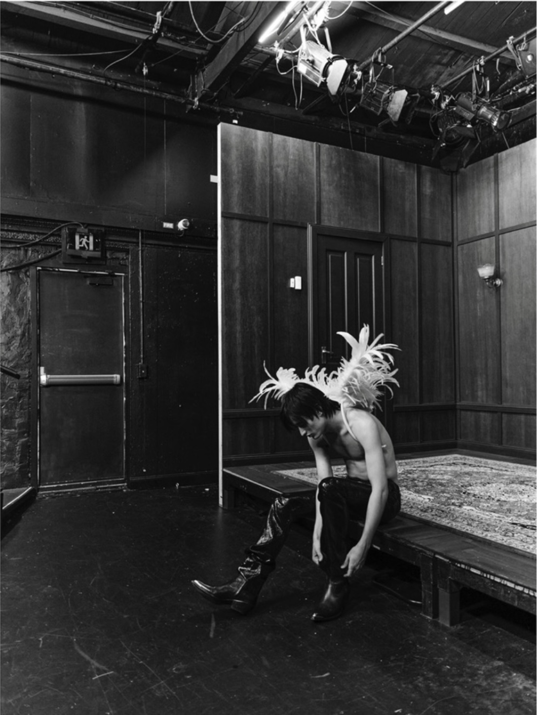 An adult male, wearing feathered shoulder pads and shiny black pants, sits on the edge of a wooden stage in a dimly lit performance space.
