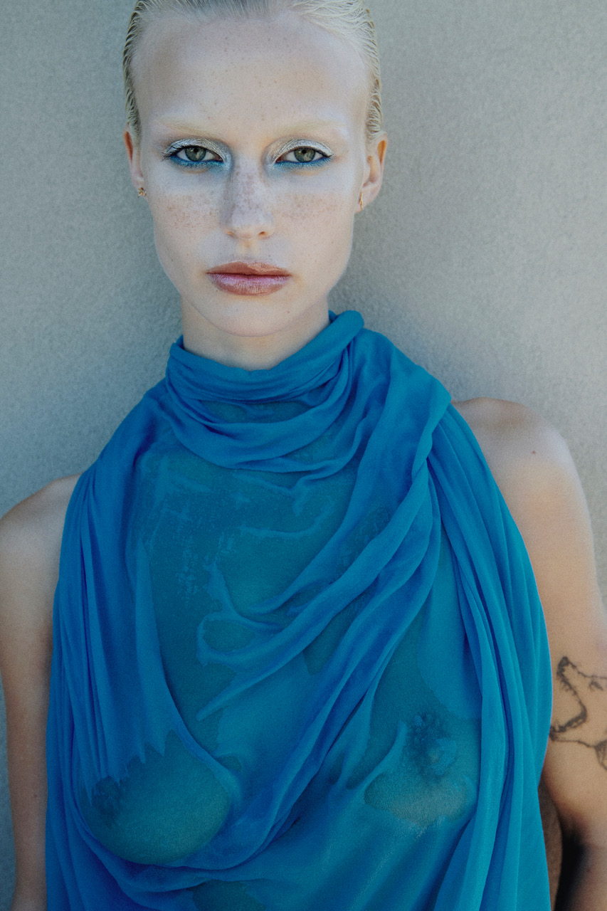 Close-up studio portrait of a fair-skinned adult female with white hair, blue eyeshadow, and a translucent blue wrap, against a neutral wall.