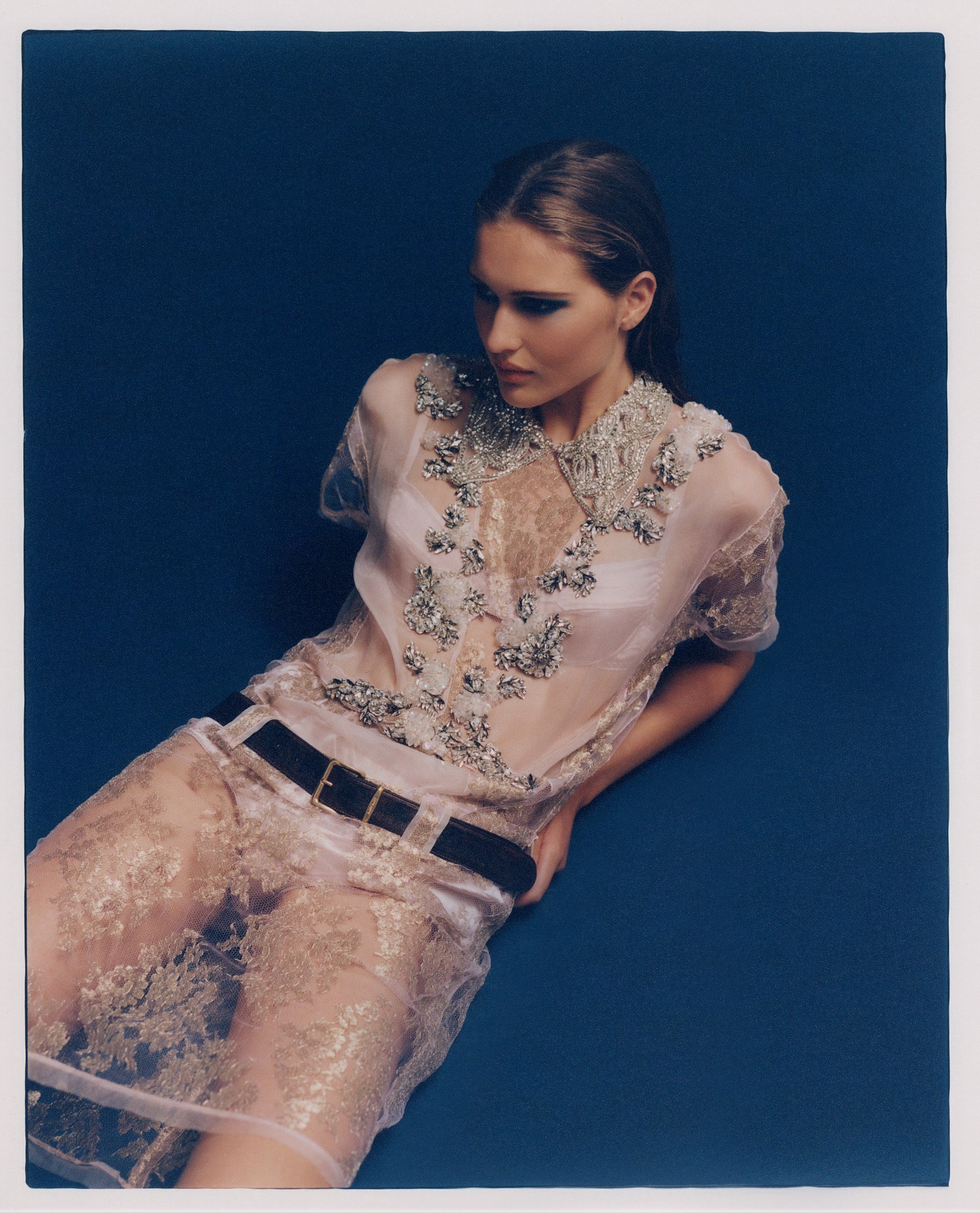 A woman poses against a blue background, wearing a sheer, embellished, short-sleeved dress with a black belt and her head angled downward.