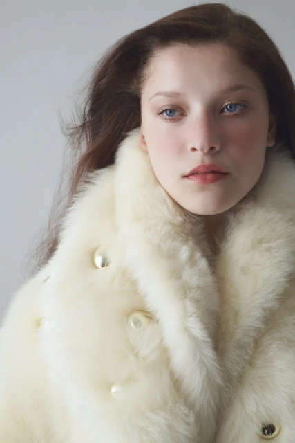 Close-up studio shot of a young woman with light skin and blue eyes wearing a white faux fur coat, looking directly at the camera.