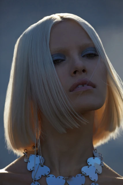 Close-up portrait of a woman with short blonde hair, wearing a silver necklace and makeup, against a blurred grey background.