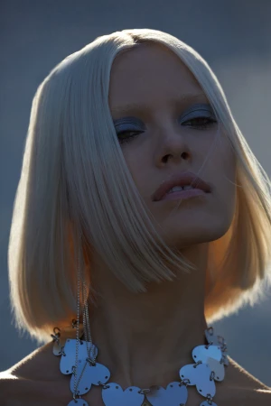 Close-up portrait of a woman with short blonde hair, wearing a silver necklace and makeup, against a blurred grey background.