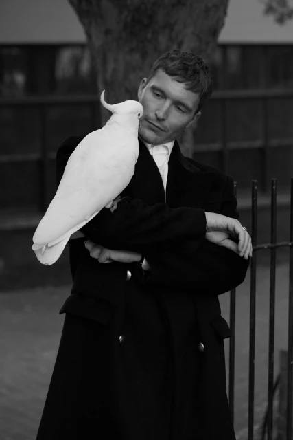 Monochrome portrait of a man in a coat with a Sulphur-crested cockatoo perched on his arm in a park setting with a metal fence.