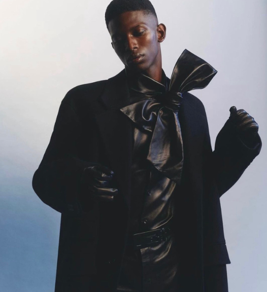 Male fashion model in black outfit featuring a large bow tie and gloves against a gradient background for editorial concept.