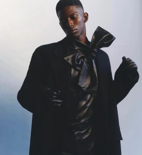 Male fashion model in black outfit featuring a large bow tie and gloves against a gradient background for editorial concept.
