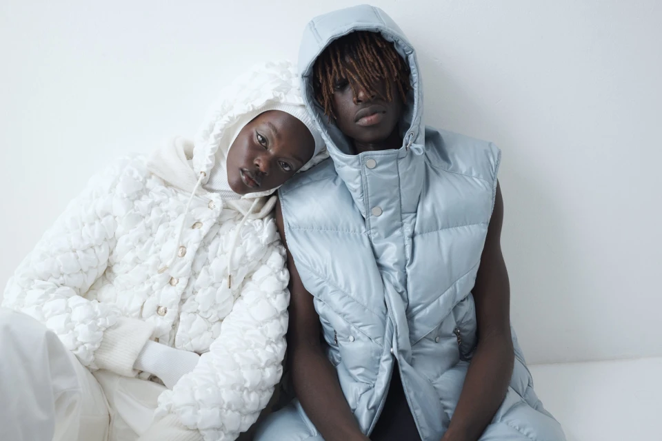 Two models pose indoors against a white wall, wearing hooded puffer jackets in a fashion editorial shot. The model on the right wears a vest.