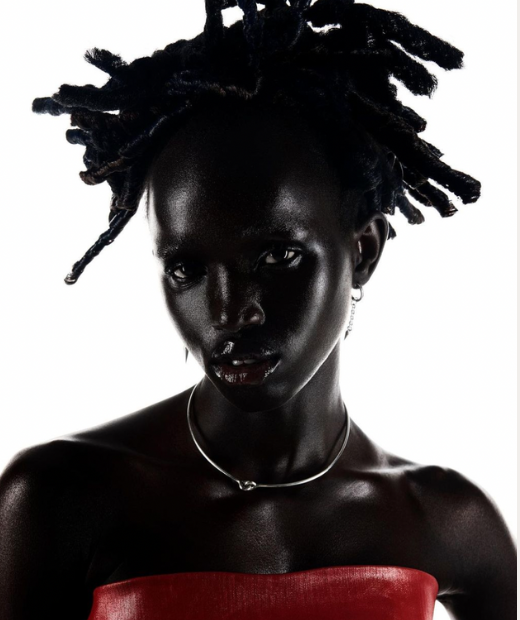 Close-up portrait of a dark-skinned woman with locs, wearing a silver choker and strapless red top, set against a bright white background.