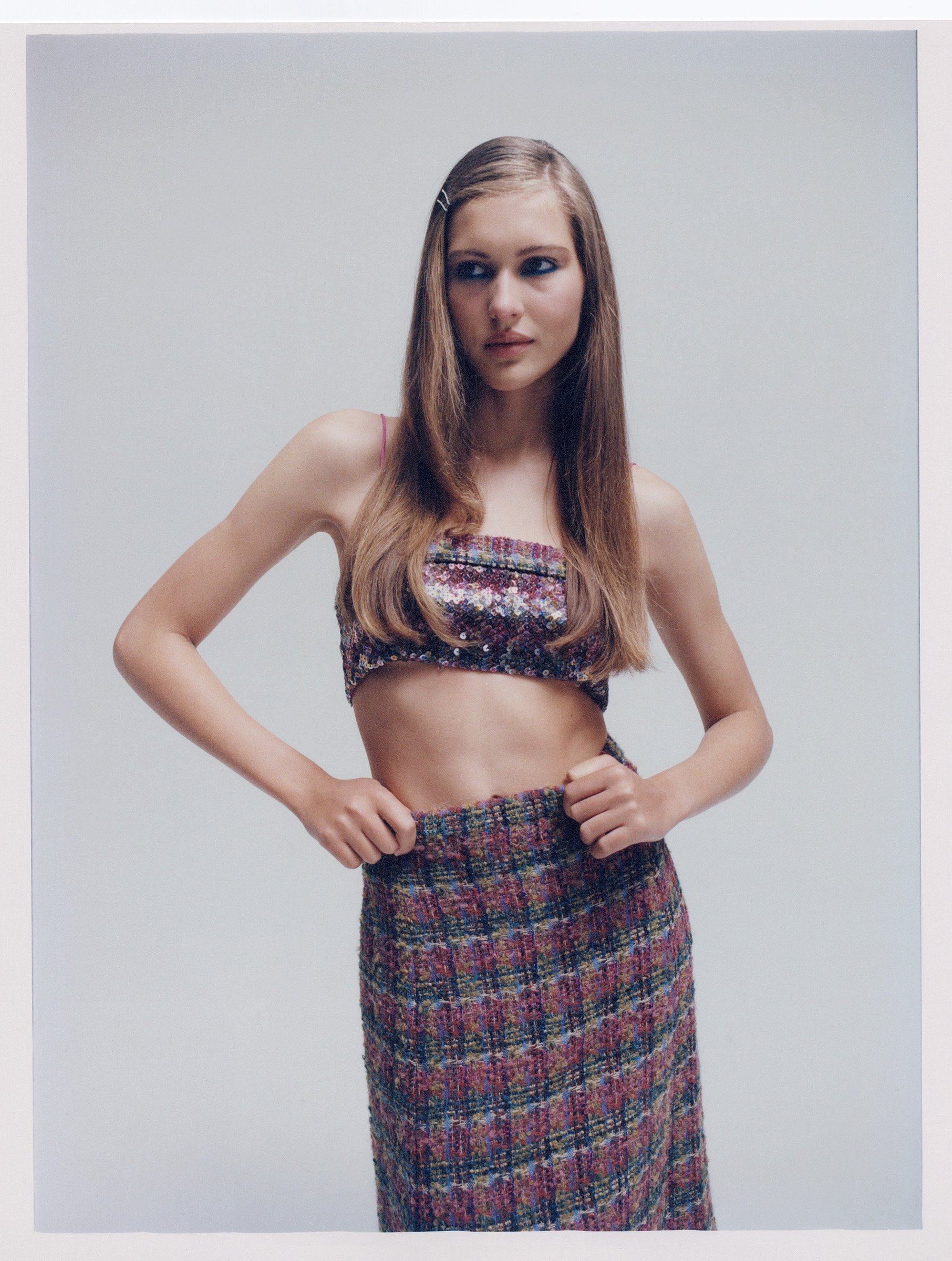 A young woman poses in a matching floral top and plaid skirt against a light gray backdrop in a studio fashion shot.