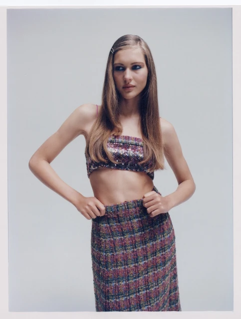 A young woman poses in a matching floral top and plaid skirt against a light gray backdrop in a studio fashion shot.