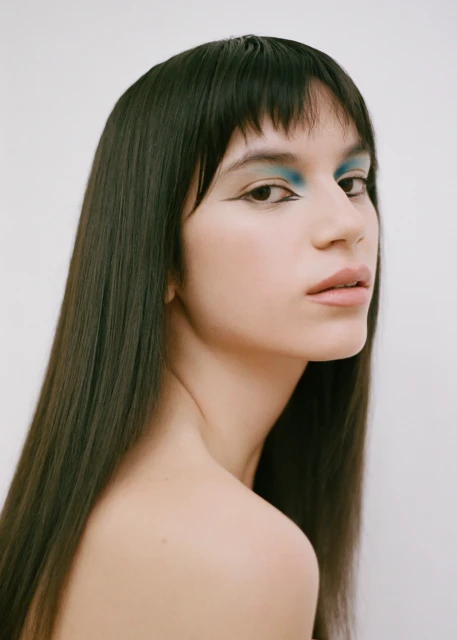 Headshot of a brunette adult female model with blue eye shadow, winged eyeliner, nude lips, bare shoulders, against a white background.