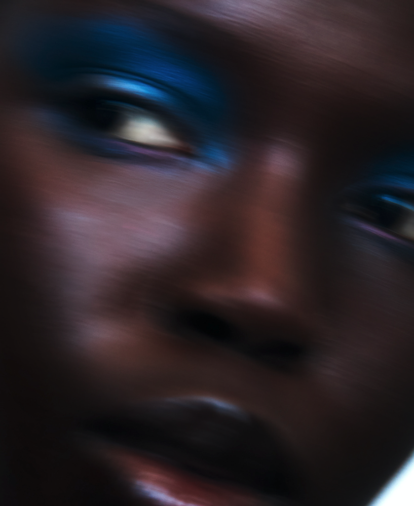 Close-up portrait of a dark-skinned woman with vibrant blue eyeshadow. The image employs a soft-focus technique for a dreamy aesthetic.