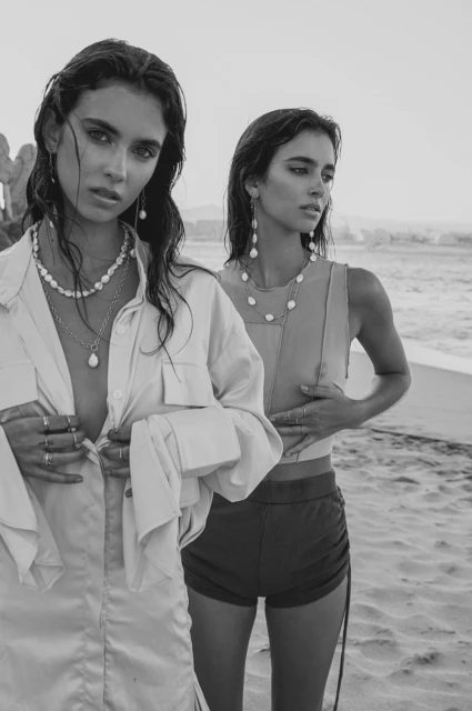 Two women pose on a beach wearing jewelry, wet hair, casual shirts and shorts in a monochromatic style with a beach background.
