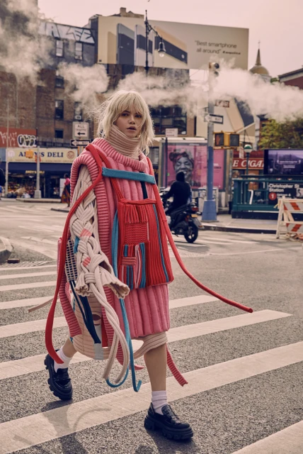 Fashionable woman in pink and blue knitwear crossing a city street with steam and buildings in the background on a sunny day.