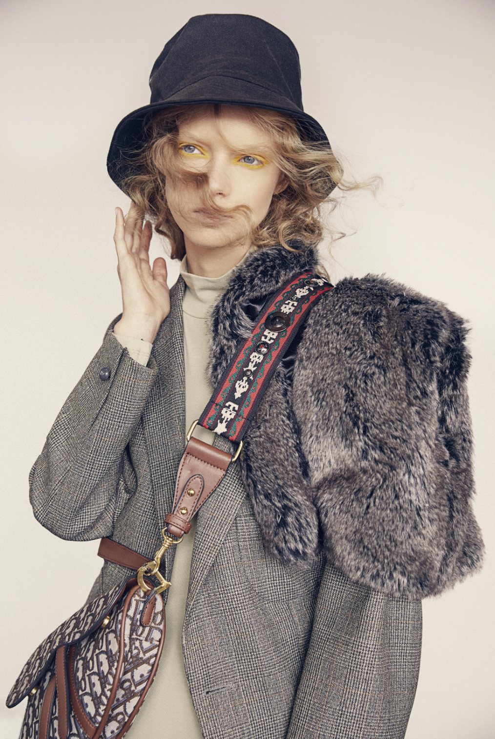 Fashion shot of a fair-skinned woman wearing a grey blazer, bucket hat, and a Dior purse with fur stole in a studio setting.