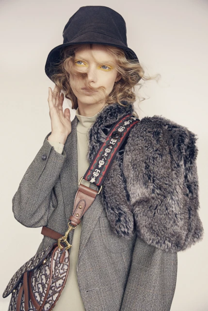 Fashion shot of a fair-skinned woman wearing a grey blazer, bucket hat, and a Dior purse with fur stole in a studio setting.