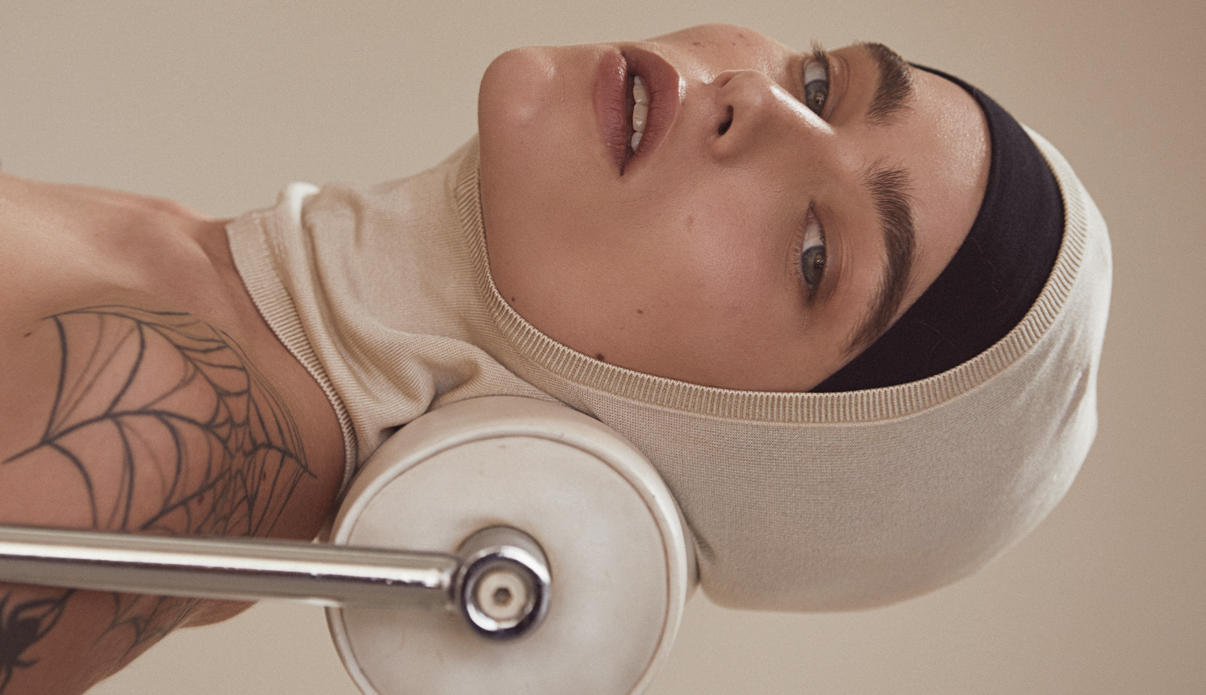 Portrait of a woman with a knit head covering and tattoo lying down against a beige background, looking up at the camera.