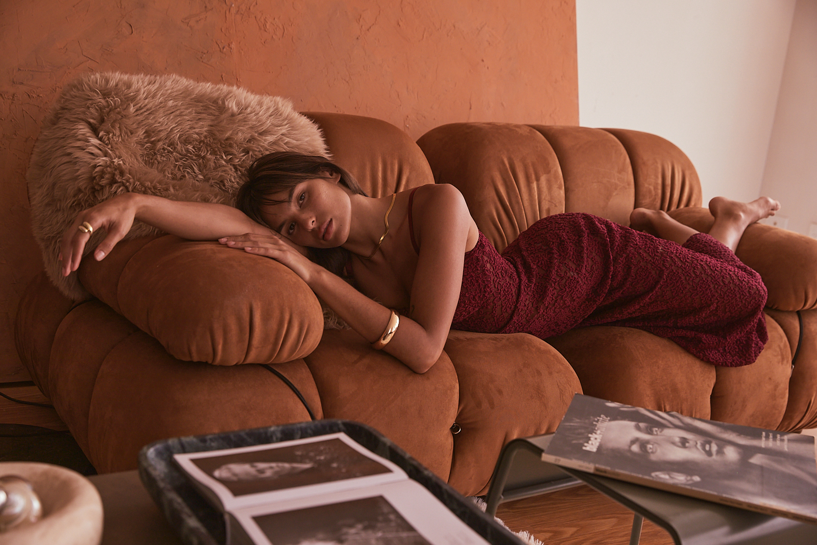 Adult woman with brown hair in a red dress reclines on a brown couch with a fur pillow in an indoor setting with magazine and decor.