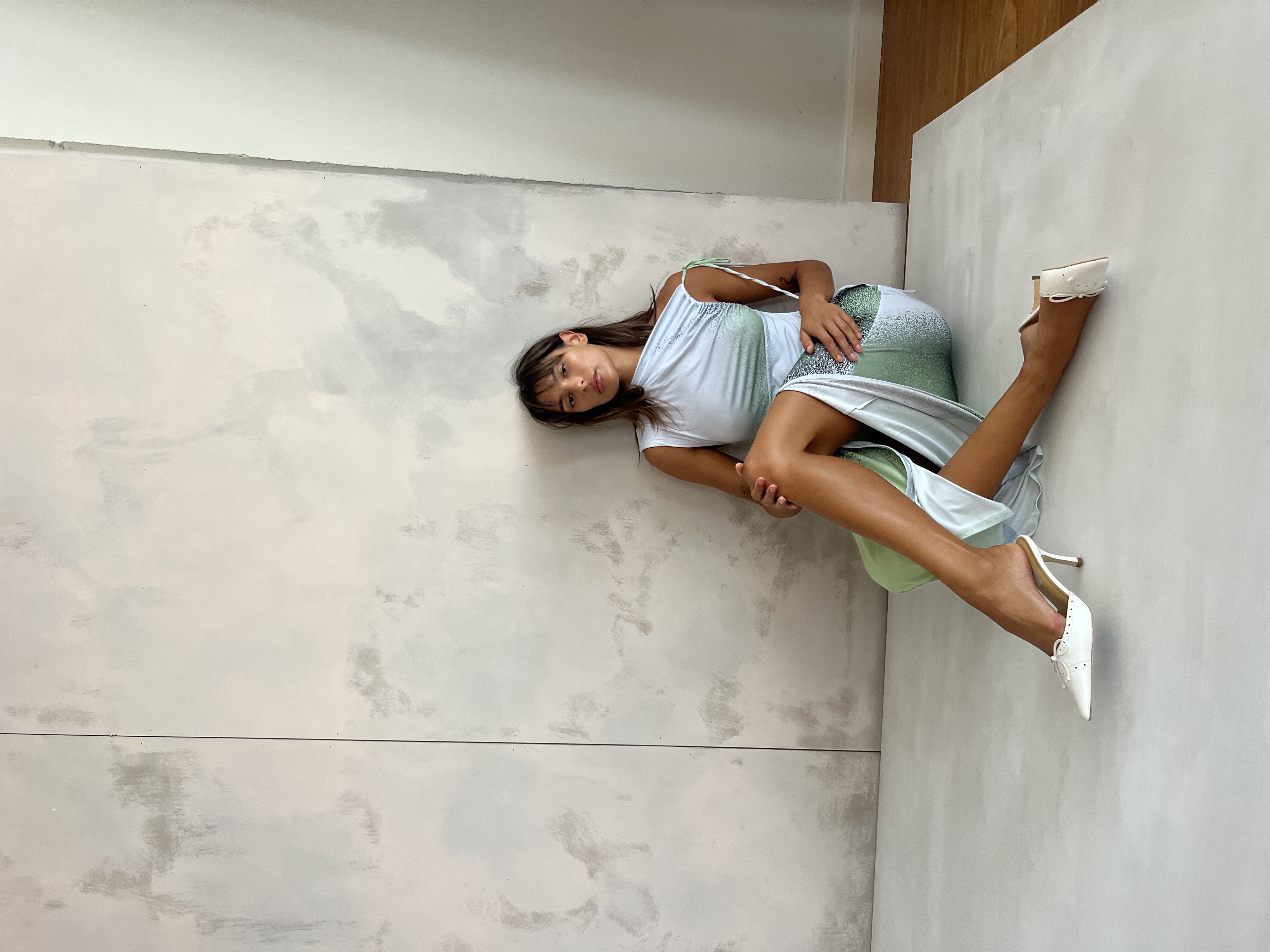 Brunette woman in a light blue dress sits on the floor in a studio setting, legs extended, wearing white heels, posing for fashion shot.