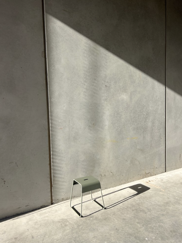 Minimalist stool stands alone on a concrete surface in sunlight, casting a sharp shadow against a stark grey concrete wall.