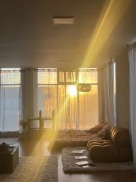 Modern living room interior with warm sunset light streaming through large windows onto a low platform bed and brown modular sofa.