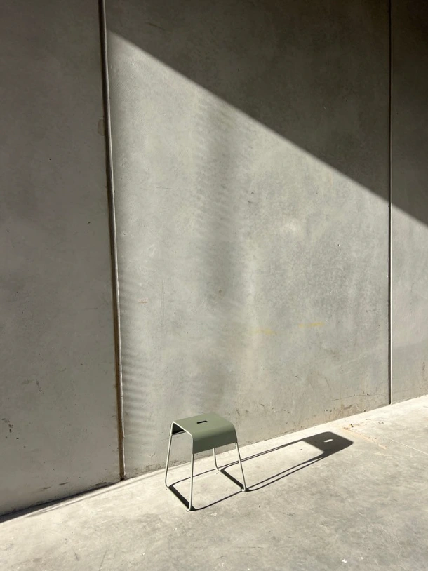 Green stool on concrete floor against a grey concrete wall with sharp diagonal sunlight and long shadows.