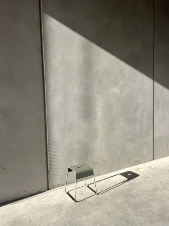Green stool on concrete floor against a grey concrete wall with sharp diagonal sunlight and long shadows.