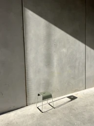 Green stool on concrete floor against a grey concrete wall with sharp diagonal sunlight and long shadows.