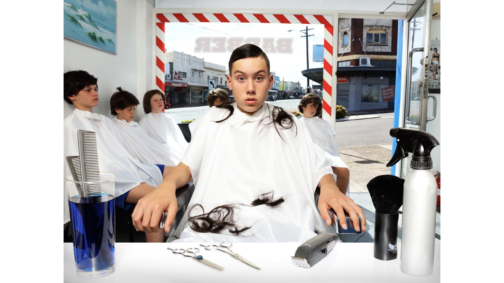 A young person sits in a barber's chair with a white cape, fresh haircut, and black hair clippings, with other clients in the background.