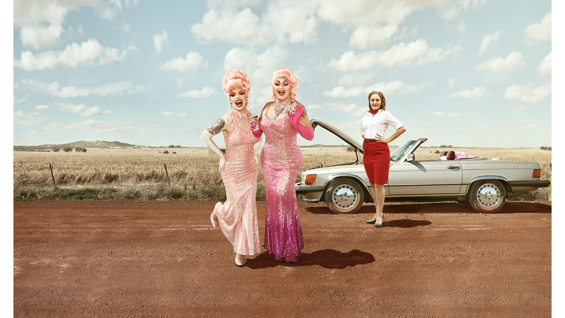 Two drag queens in pink gowns pose on a dirt road next to a classic convertible, with a woman standing by the open hood.