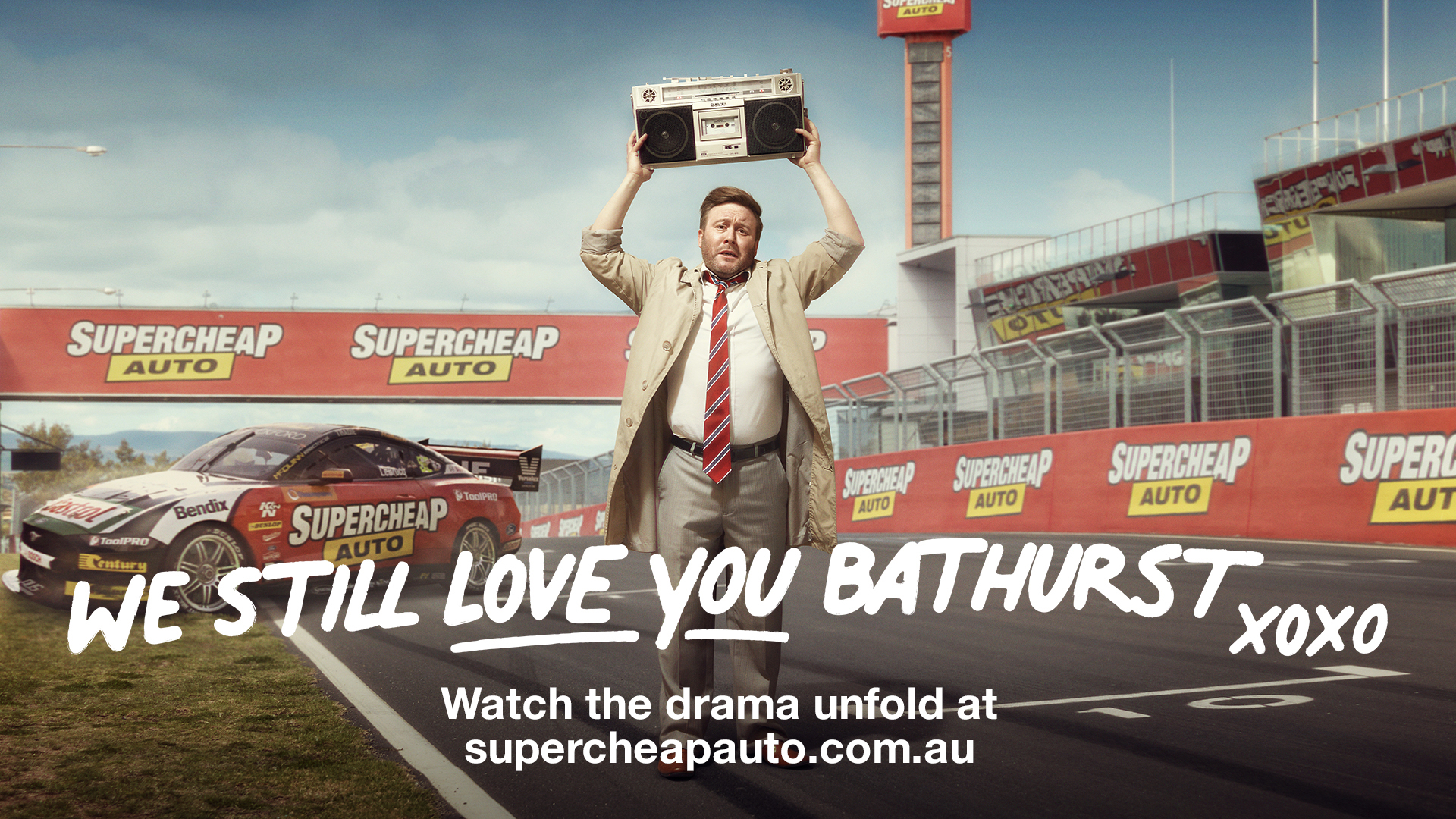 Promotional shot for Supercheap Auto featuring a man holding a boombox above his head with race car and track in the background.