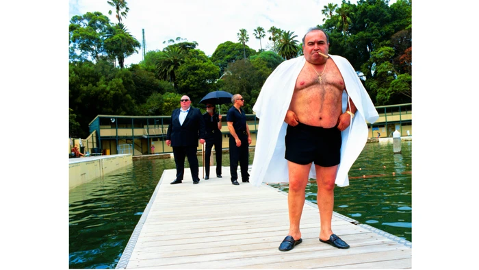 A man wearing a white cape over black shorts holding a cigar stands on a wooden pier with three men in the background by Neil Bailey.