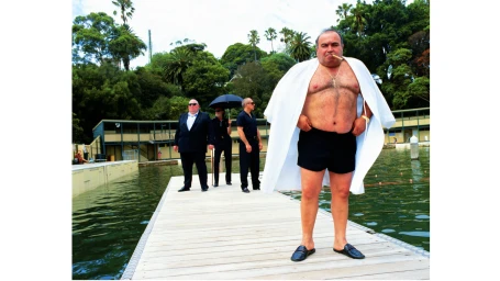 A man wearing a white cape over black shorts holding a cigar stands on a wooden pier with three men in the background by Neil Bailey.