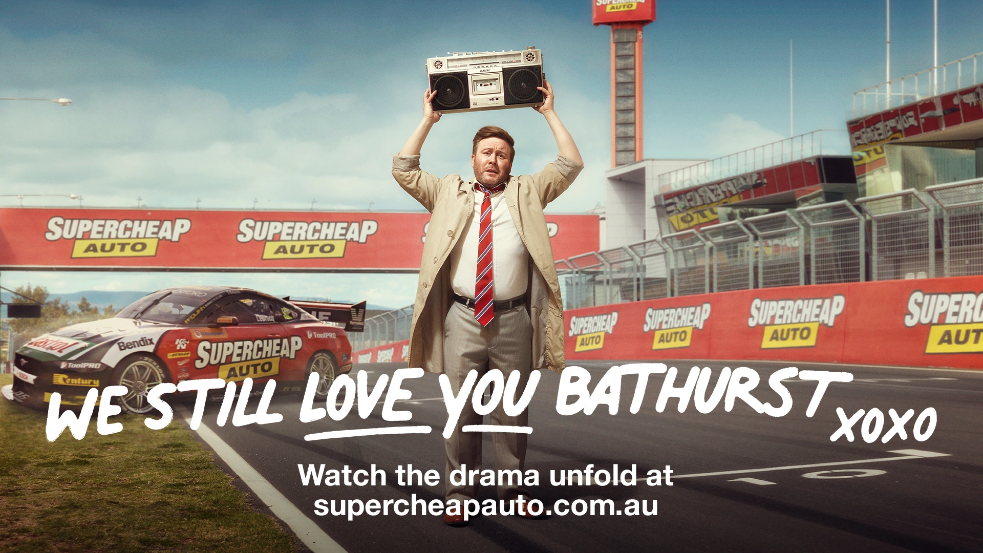 Man holding a boombox above his head on a racetrack holding red and white barriers with a race car parked nearby.