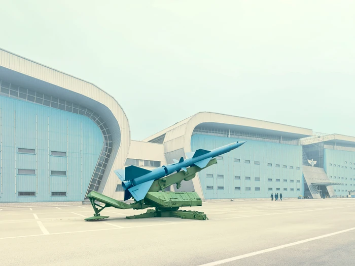 Blue and green missile launcher parked outside large modern industrial buildings under a pale sky, photographed by Scott Newett