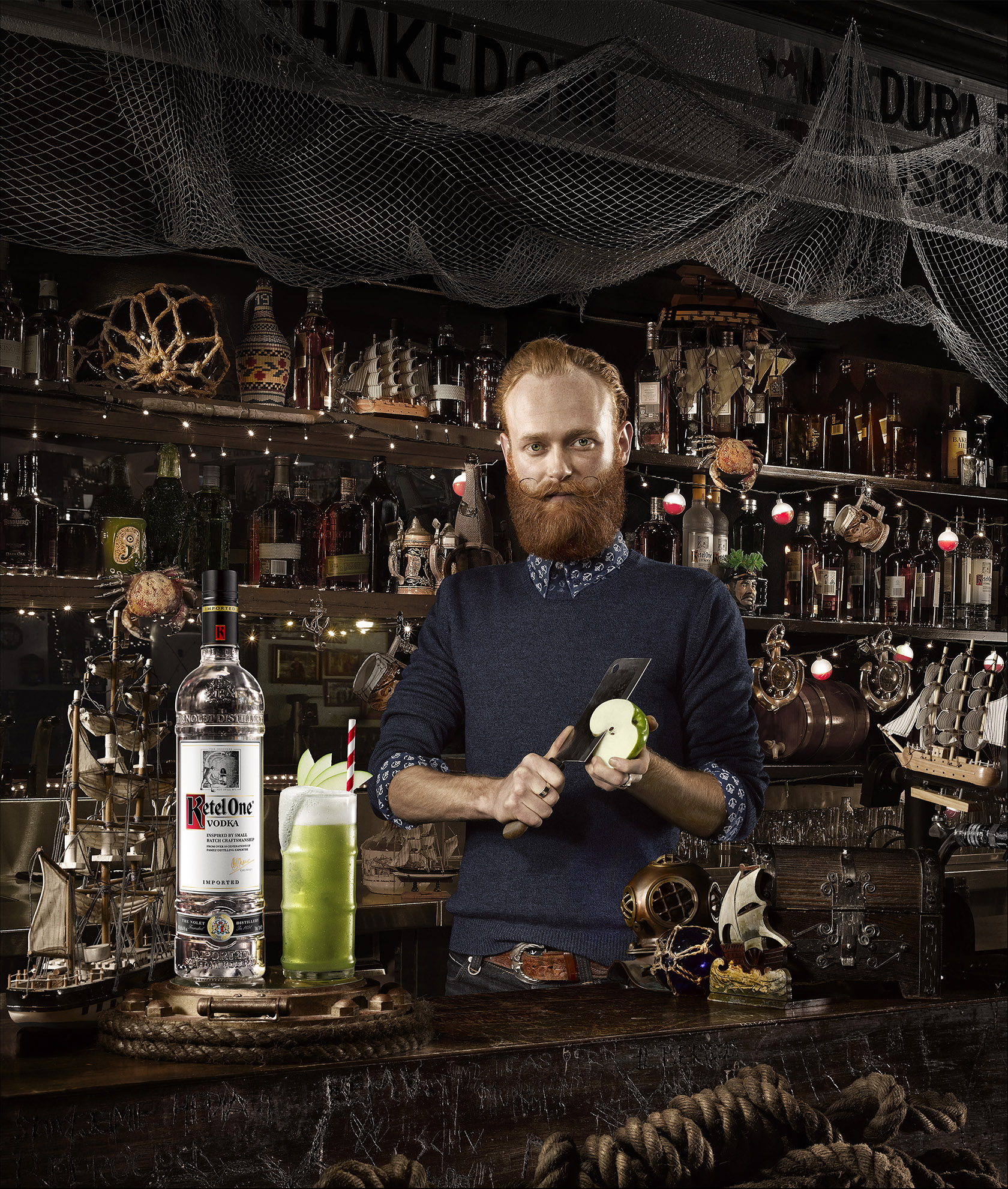 Person preparing cocktail with apple and liquor bottle on bar with nautical decorations and bar lighting in the background.