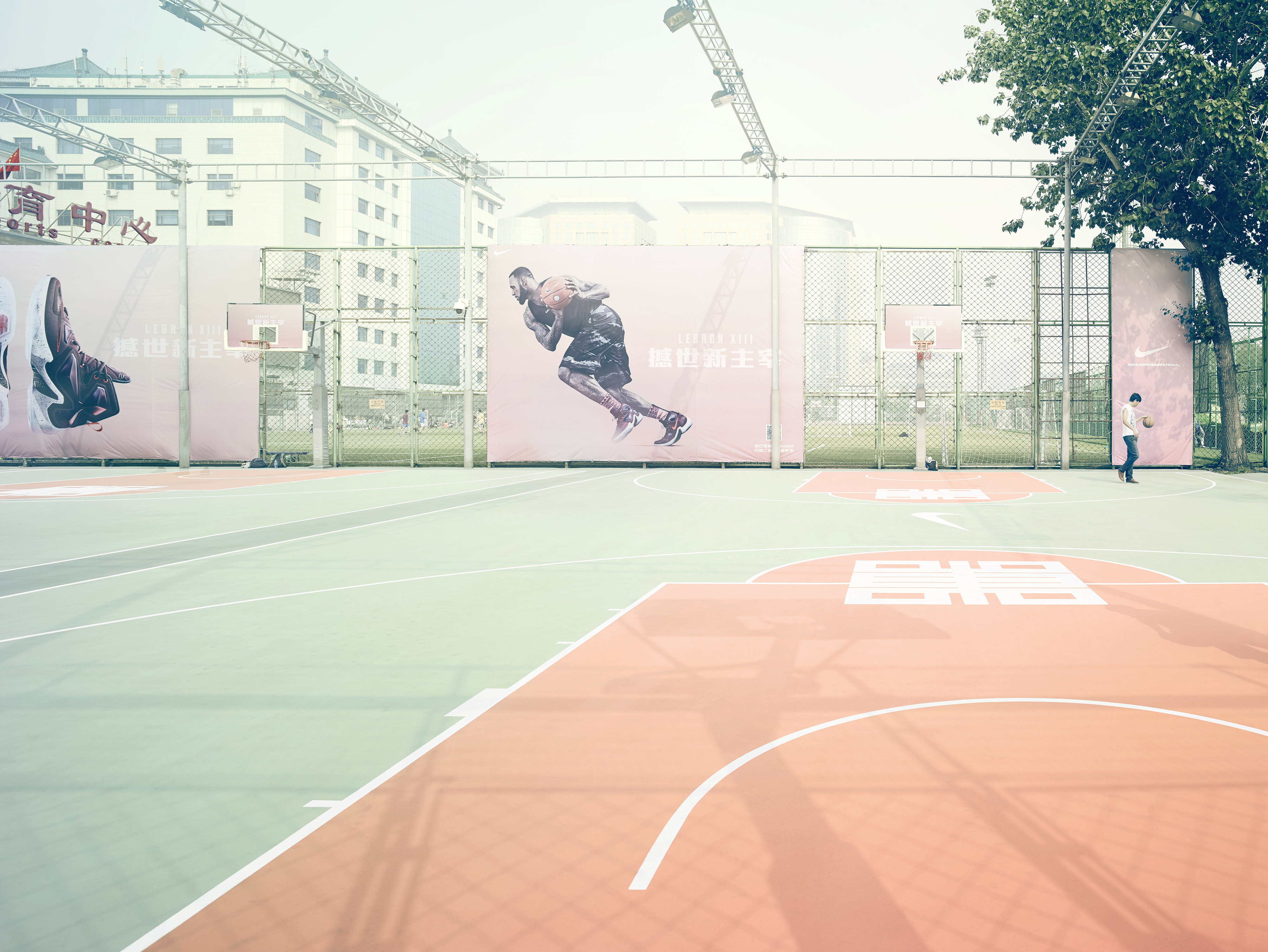 Outdoor basketball court featuring a basketball hoop, a person holding a ball, and Nike advertising posters in the background, daytime.