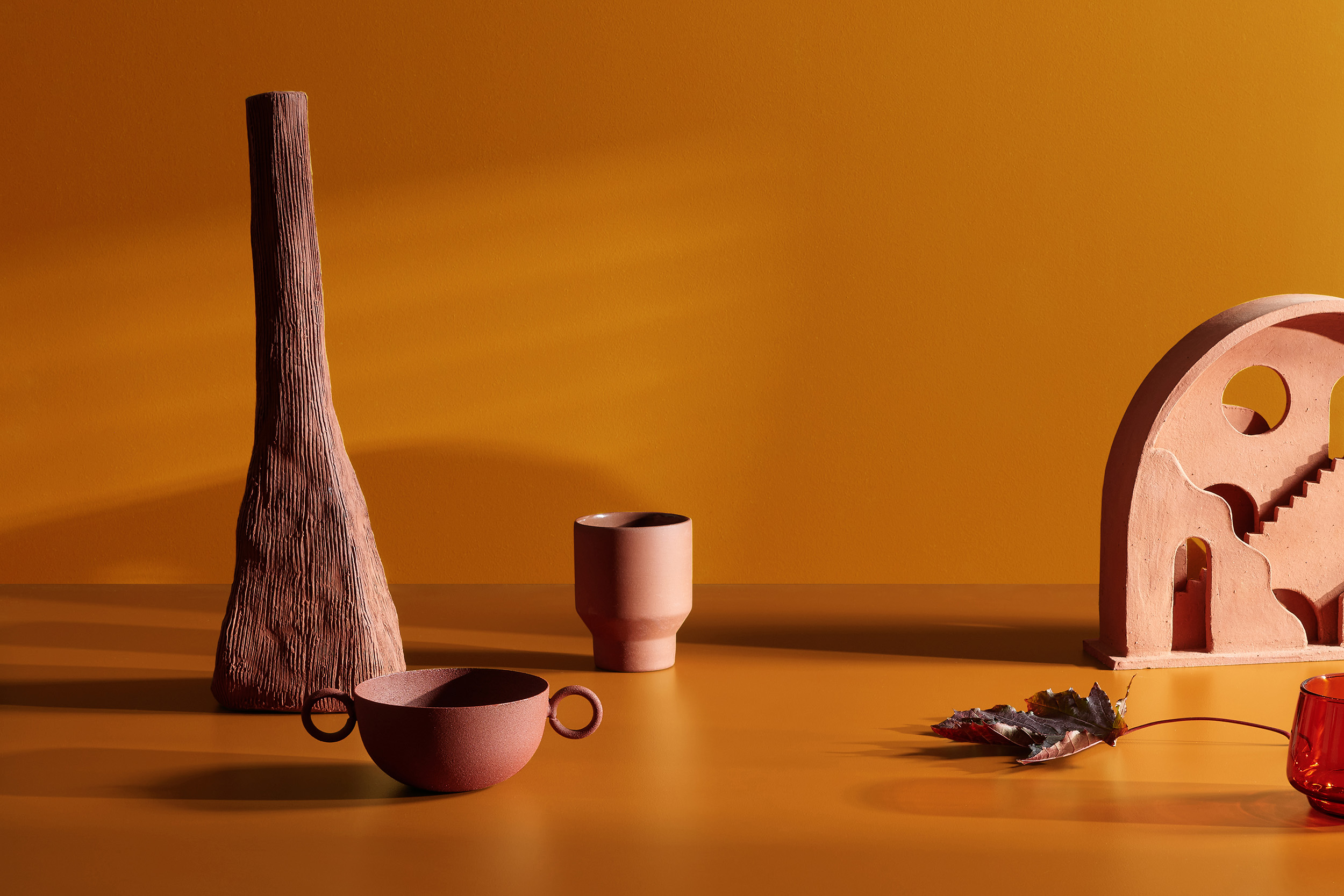 Still life of terracotta vases, cups, and a decorative archway on an orange surface against a warm orange background.