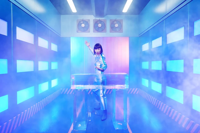 Woman in futuristic suit poses behind ice block in blue-lit room with fans, in a scene with dynamic lighting and stage-like atmosphere.