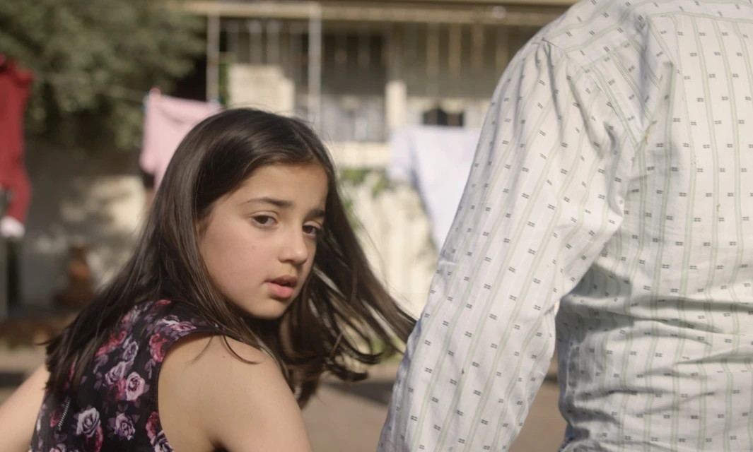 Girl with long brown hair stands next to an adult in a patterned shirt, in an outdoor, sunlit environment with hanging laundry in the background.