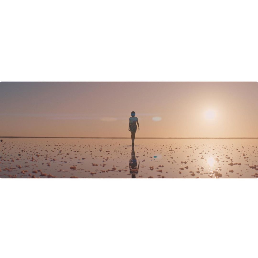 A single adult walking across a large, reflective salt flat at sunset, with the sun low on the horizon and the sky tinted with orange and pink.