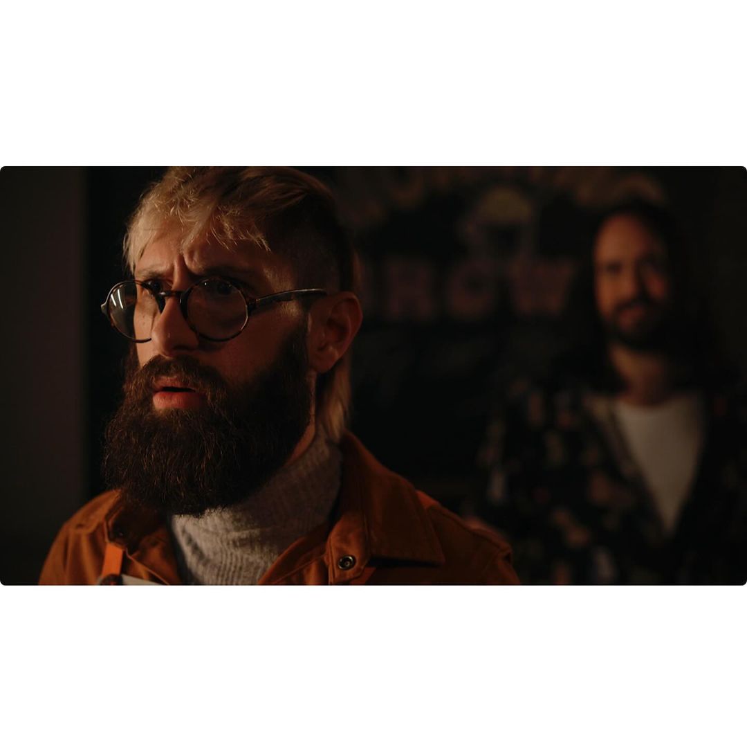 Two men in a dimly lit indoor setting. A bearded man with glasses is in the foreground, while a second man stands blurred in the background.