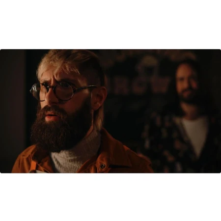 Two men in a dimly lit indoor setting. A bearded man with glasses is in the foreground, while a second man stands blurred in the background.